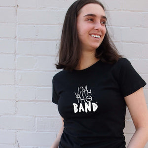 I'm With The Band® white logo print on black close-fit women's T-shirt on cool young woman standing in front of a painted brick wall