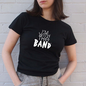 I'm With The Band® white logo print on black close-fit women's T-shirt on cool young woman who is facing at the camera although we can just see her lips and standing in front of a painted brick wall