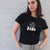 I'm With The Band® white logo print on black close-fit women's T-shirt on cool young woman who is looking at the camera and standing in front of a painted brick wall