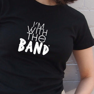 I'm With The Band® white logo print on black close-fit women's T-shirt very close up view on cool young woman facing the camera and standing in front of a painted brick wall