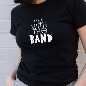 I'm With The Band® white logo print on black close-fit women's T-shirt very close up view on cool young woman standing in front of a painted brick wall