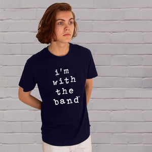 I'm With The Band® Wonky typewriter logo graphic with white print on navy T being worn by a cool young man standing against a white painted brick wall and looking into the distance.