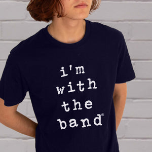 Close up of I'm With The Band® Wonky typewriter logo graphic with white print on navy T being worn by a cool young man standing against a white painted brick wall.