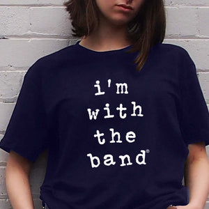 Close up of I'm With The Band® Wonky typewriter logo graphic with white print on navy T being worn by a cool young woman standing against a white painted brick wall.