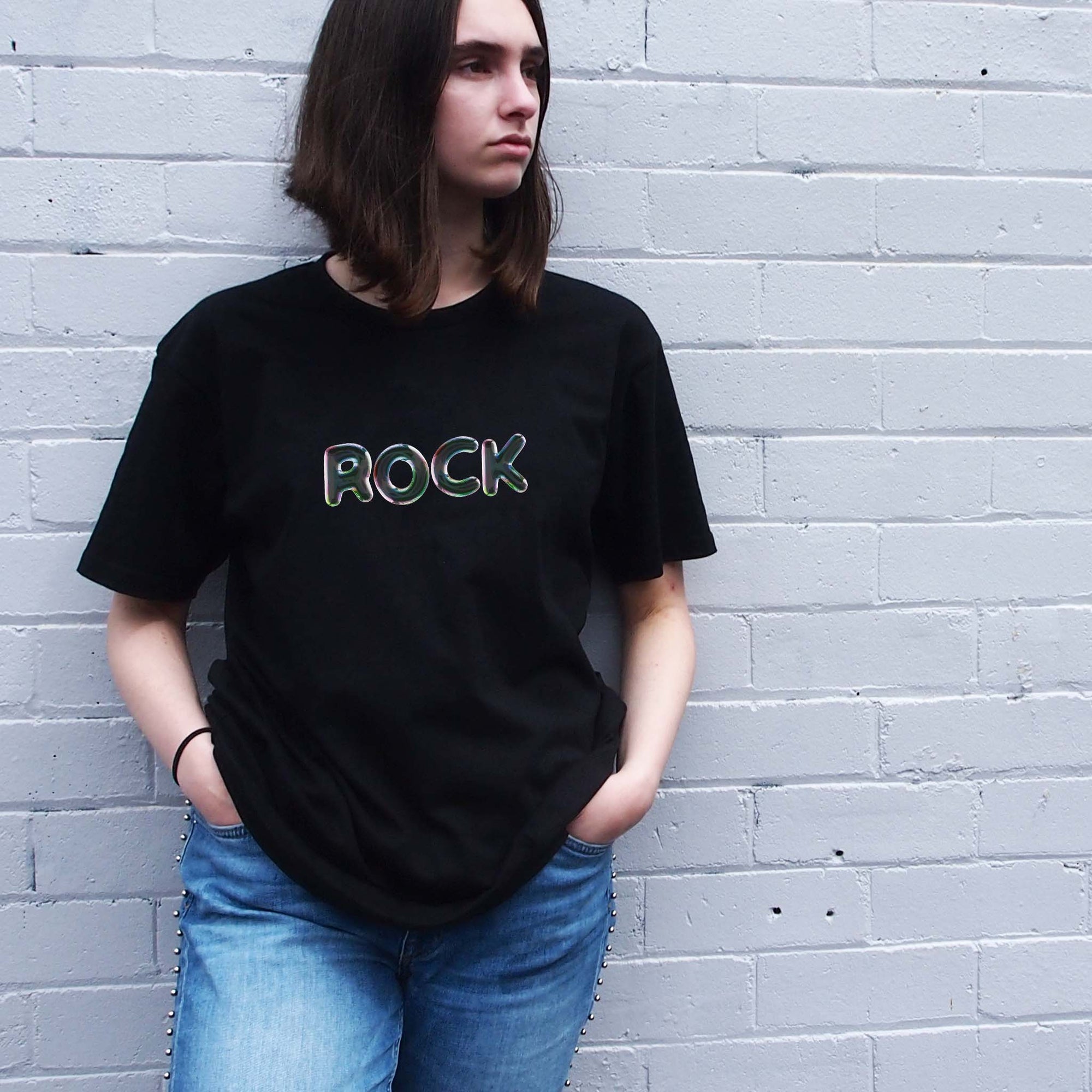 I'm With The Band® Bubble Rock T-shirt with iridescent oil slick grey print on black T being worn by a cool young woman standing against a white painted brick wall