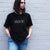 I'm With The Band® Bubble Rock T-shirt with iridescent oil slick grey print on black T being worn by a cool young woman standing against a white painted brick wall