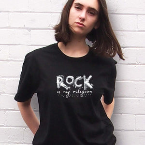 Rock Is My Religion T-shirt byI'm With The Band® with original hand-painted white graphic print on black T being worn by a cool young woman who is looking into the camera and standing against a white painted brick wall