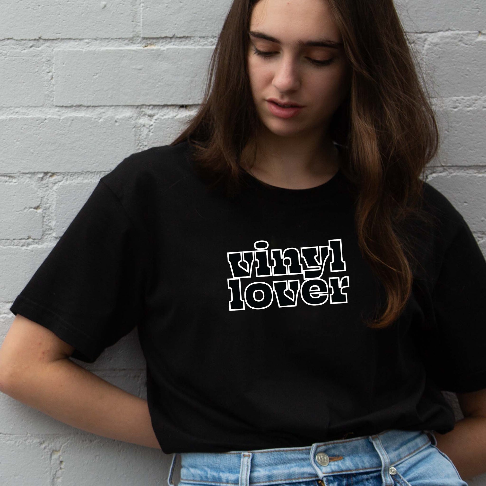 I'm With The Band® Vinyl Lover T-shirt with black print white outline on black T being worn by a cool young woman leaning against a white painted brick wall