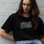 I'm With The Band® Vinyl Lover T-shirt with black print white outline on black T being worn by a cool young woman leaning against a white painted brick wall