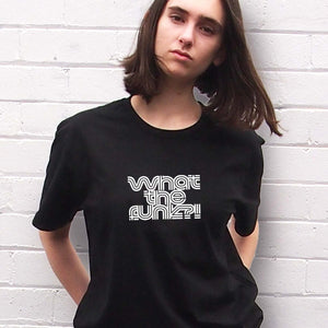 I'm With The Band® What The Funk T-shirt with white print on black T  on a cool young woman looking at the camera and standing in front of a painted brick wall